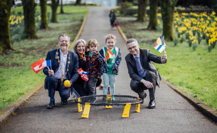 Two men crouched down, a mayor and minister, with a mother and two young children holding european flags in a city park with the ball and bet from a game of spike ball in front of them