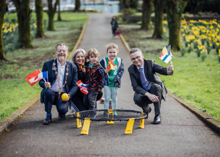 Two men crouched down, a mayor and minister, with a mother and two young children holding european flags in a city park with the ball and bet from a game of spike ball in front of them