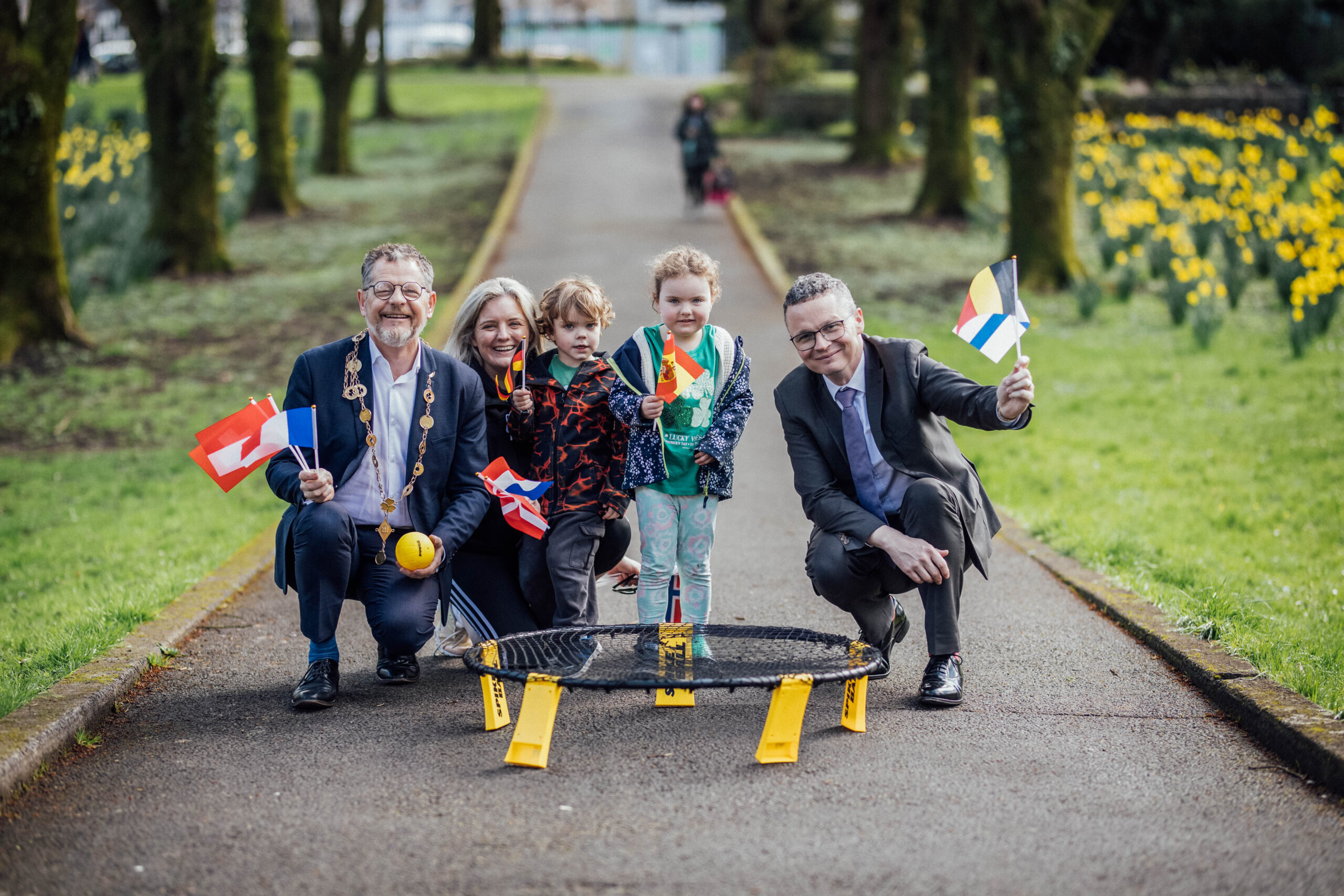Limerick European Sport for All Launch 032 Two men crouched down, a mayor and minister, with a mother and two young children holding european flags in a city park with the ball and bet from a game of spike ball in front of them