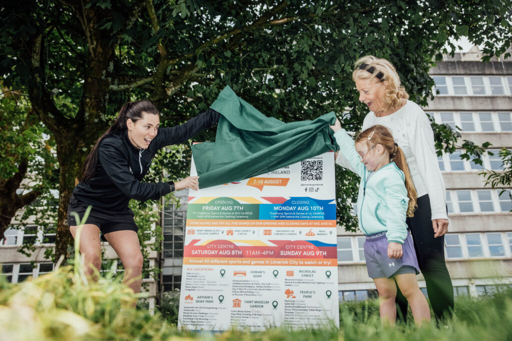 a woman unveils a large poster sign of festival programme details to a child and her mother in a park