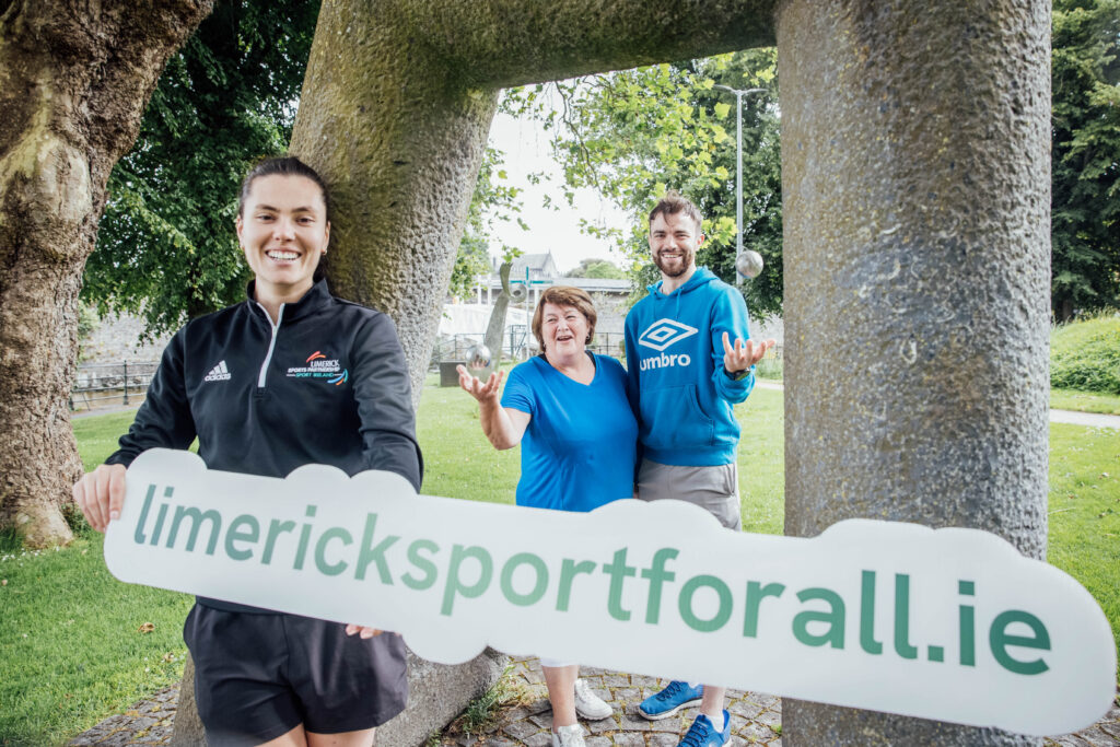 girl in a park holding cutout of website address limericksportforall.ie with a woman and man in the background