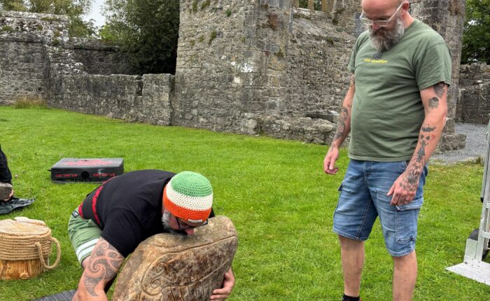 two men attempt to life a large stone in a green outdoor area