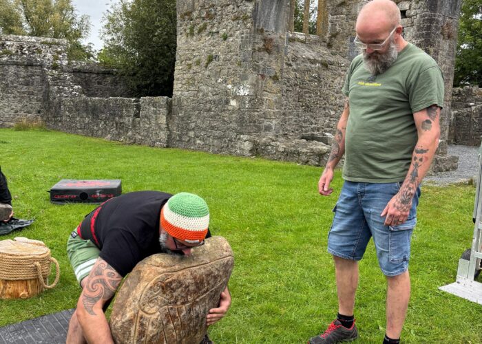 two men attempt to life a large stone in a green outdoor area