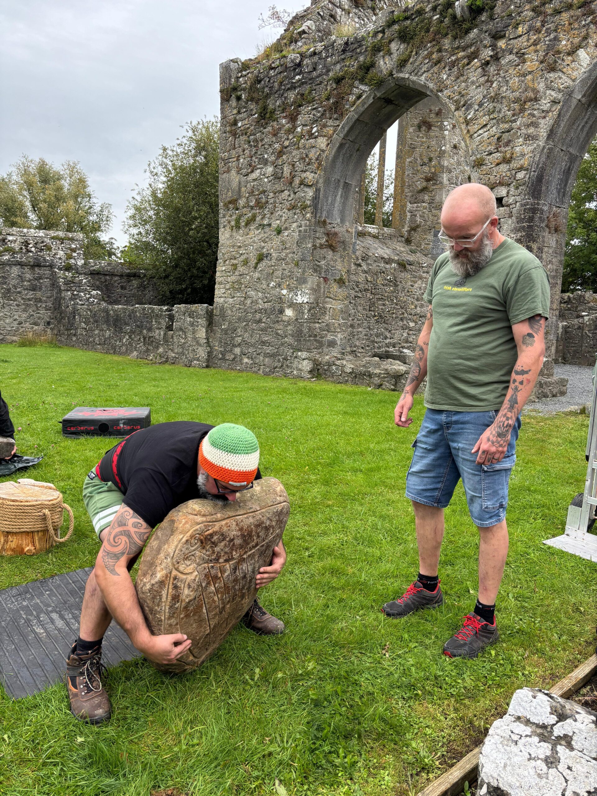 two men attempt to life a large stone in a green outdoor area